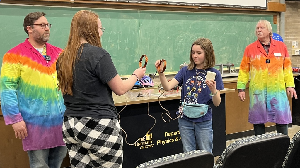 A photo of two kids taking part in a physics demonstration show with two scientists in tye-dyed lab coats standing nearby.