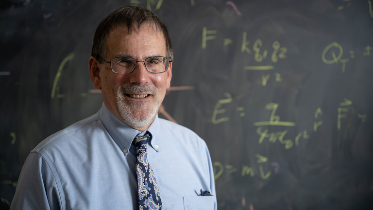 A photograph of Craig Kletzing in front of a black chalkboard with equations written.
